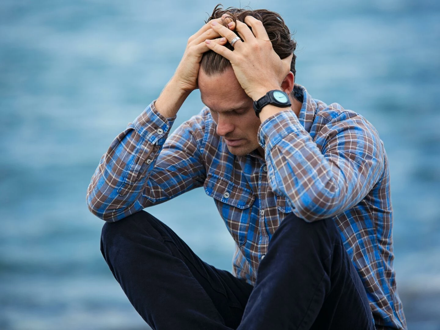 a man in a plaid shirt sits by the water looking distressed symbolizing stress