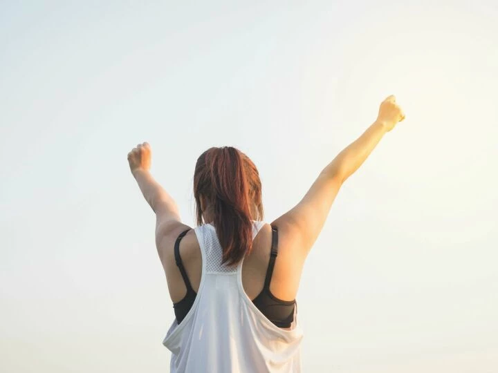 A woman celebrates success with arms raised in a bright outdoor setting.