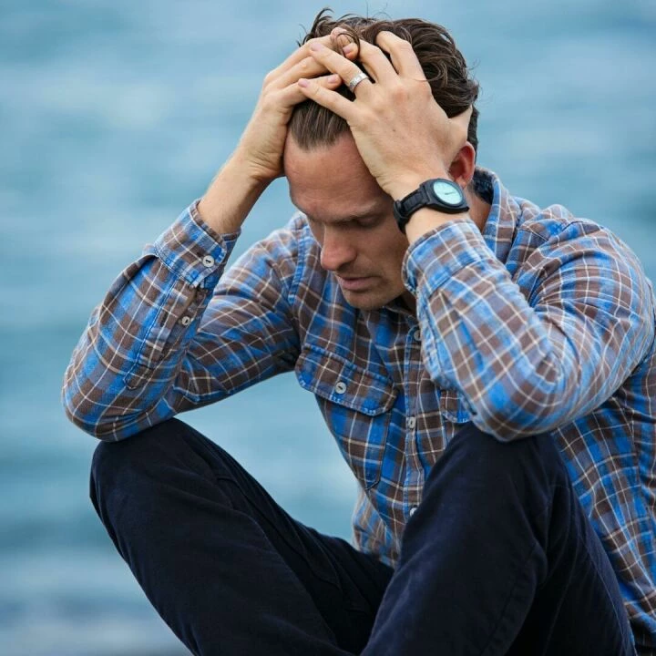 A man in a plaid shirt sits by the water looking distressed, symbolizing stress.